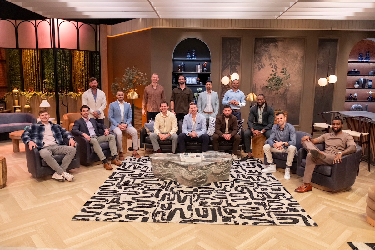 A group of 15 men sit and stand in a stylish, modern lounge with warm lighting, decorative shelves, indoor plants, and abstract art. They pose around a stone coffee table on a patterned rug, creating a relaxed, casual atmosphere.