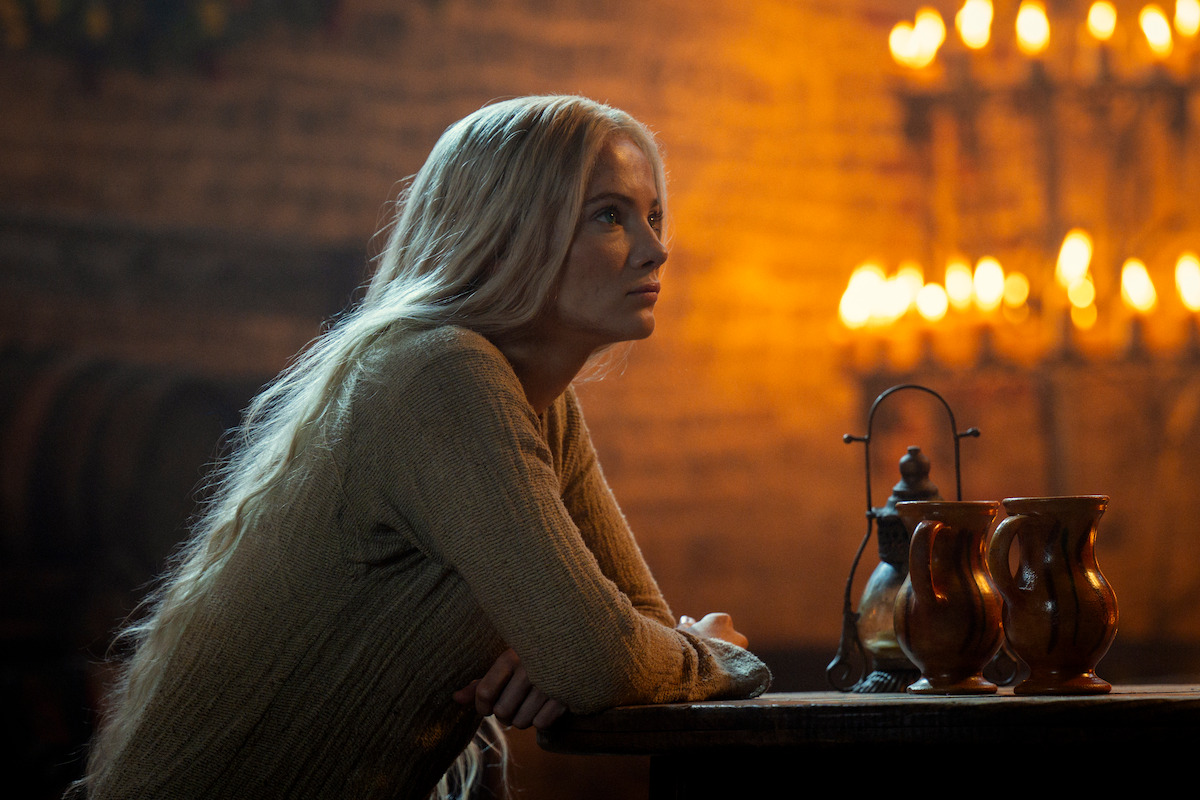 A woman with long blonde hair sits at a wooden table in a dimly lit room with candles on the wall, creating a warm and contemplative atmosphere.
