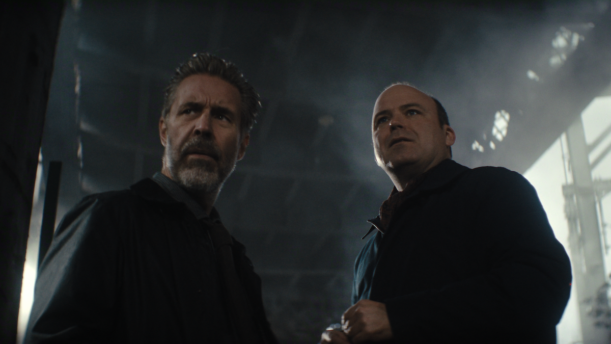 Two serious men stand indoors in a dimly lit, industrial-looking space with broken ceiling panels and dramatic lighting, both looking slightly upward with concerned expressions.