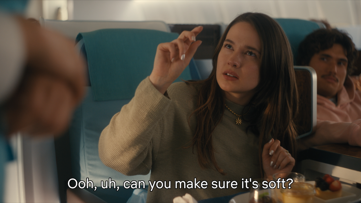 Woman sitting in an airplane seat gestures with her hand and speaks to a flight attendant, asking, "Ooh, uh, can you make sure it's soft?" Other passengers are seated nearby, with airplane interior visible.