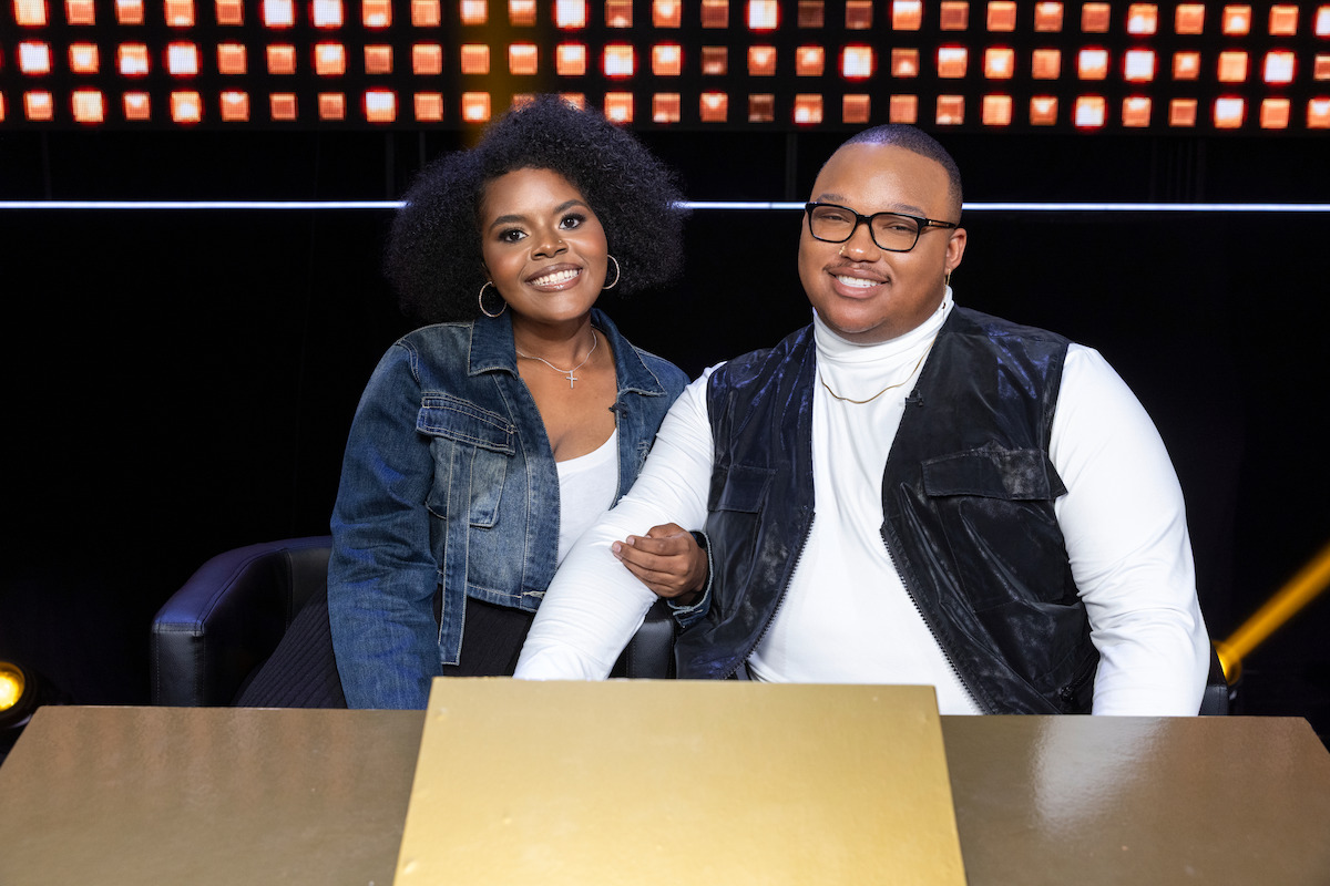 Two people sitting together at a table on a brightly lit game show set, smiling and appearing confident, with a warm and upbeat atmosphere.