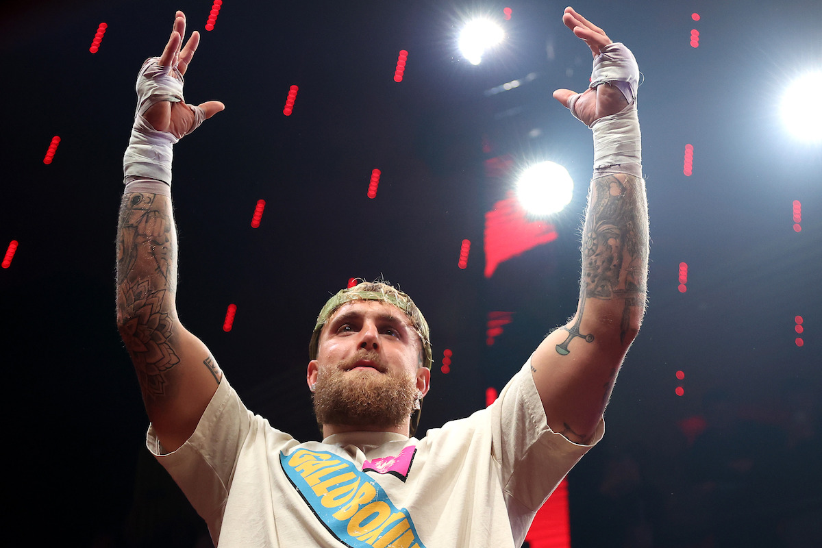 Jake Paul with bandaged hands raises arms in victory inside a brightly lit arena, wearing a t-shirt and headband, with tattoos visible on both arms and red stage lights in the background.