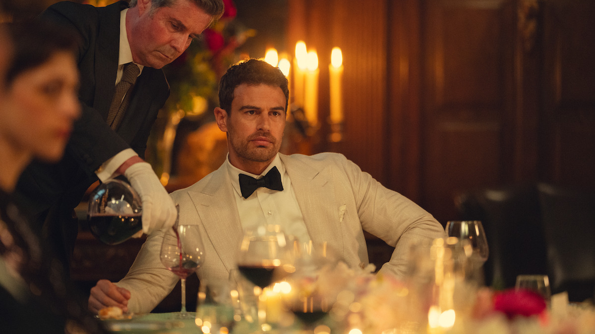 A man in a white tuxedo sits at an elegant candlelit dinner table, with a server pouring wine beside him. The setting is formal, with wine glasses, flowers, and blurred guests in the background, suggesting a sophisticated event.