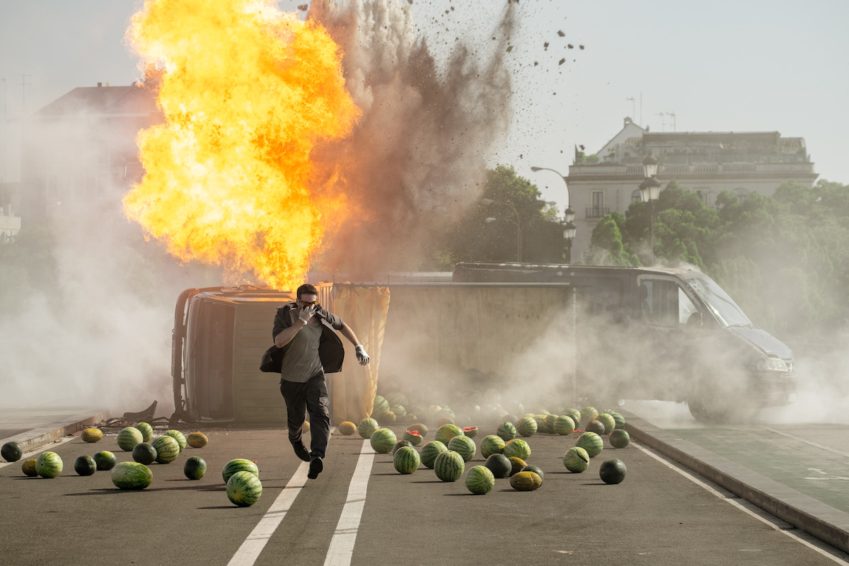 A man in a suit runs from an exploding overturned van on a city street scattered with watermelons, with smoke and fire in the background and buildings visible beyond the chaos.