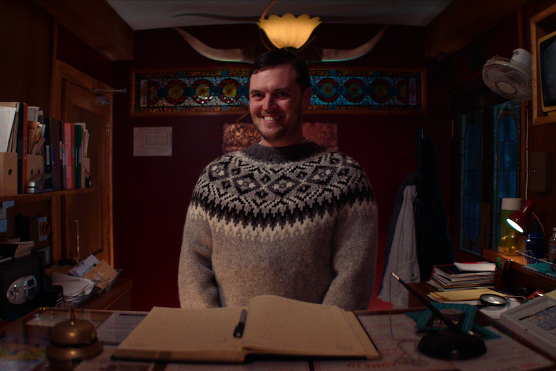 Man in patterned sweater stands behind reception desk in warmly lit, cozy office with books, folders, decorative stained glass, and antler decor above. Open ledger and vintage-style items are on the desk in front of him.