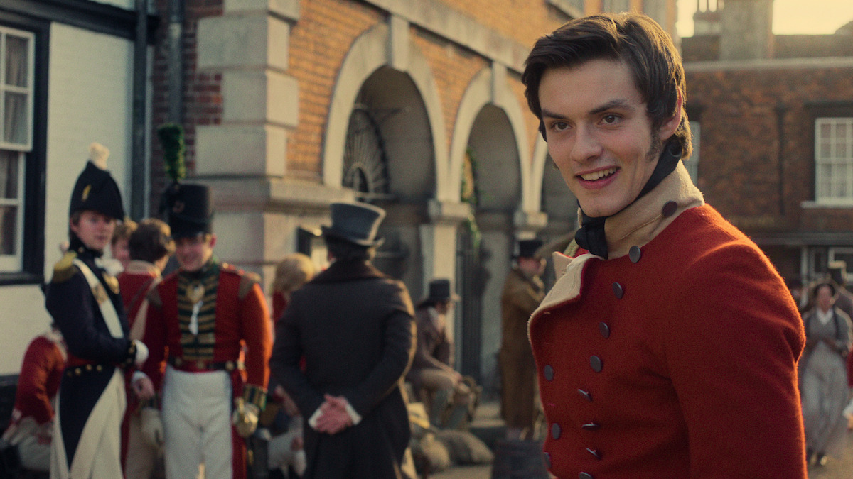 Young man in red historical military uniform smiles in foreground, with other soldiers and townspeople gathered outside a brick building with arches in a period village setting, evoking a historical or Regency-era atmosphere.