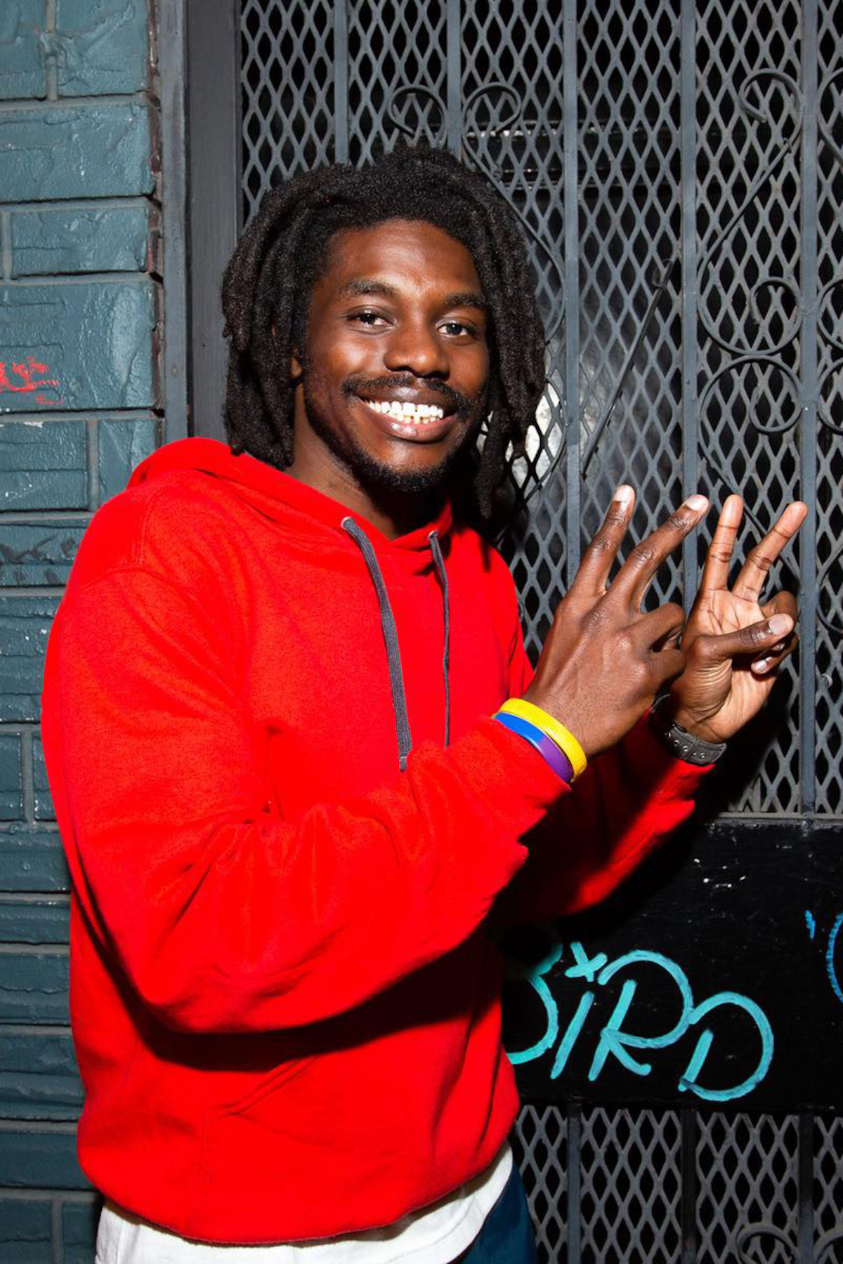 Smiling man in a red hoodie with wristbands making peace signs, standing in front of a metal gate and graffiti-covered brick wall in an urban setting.