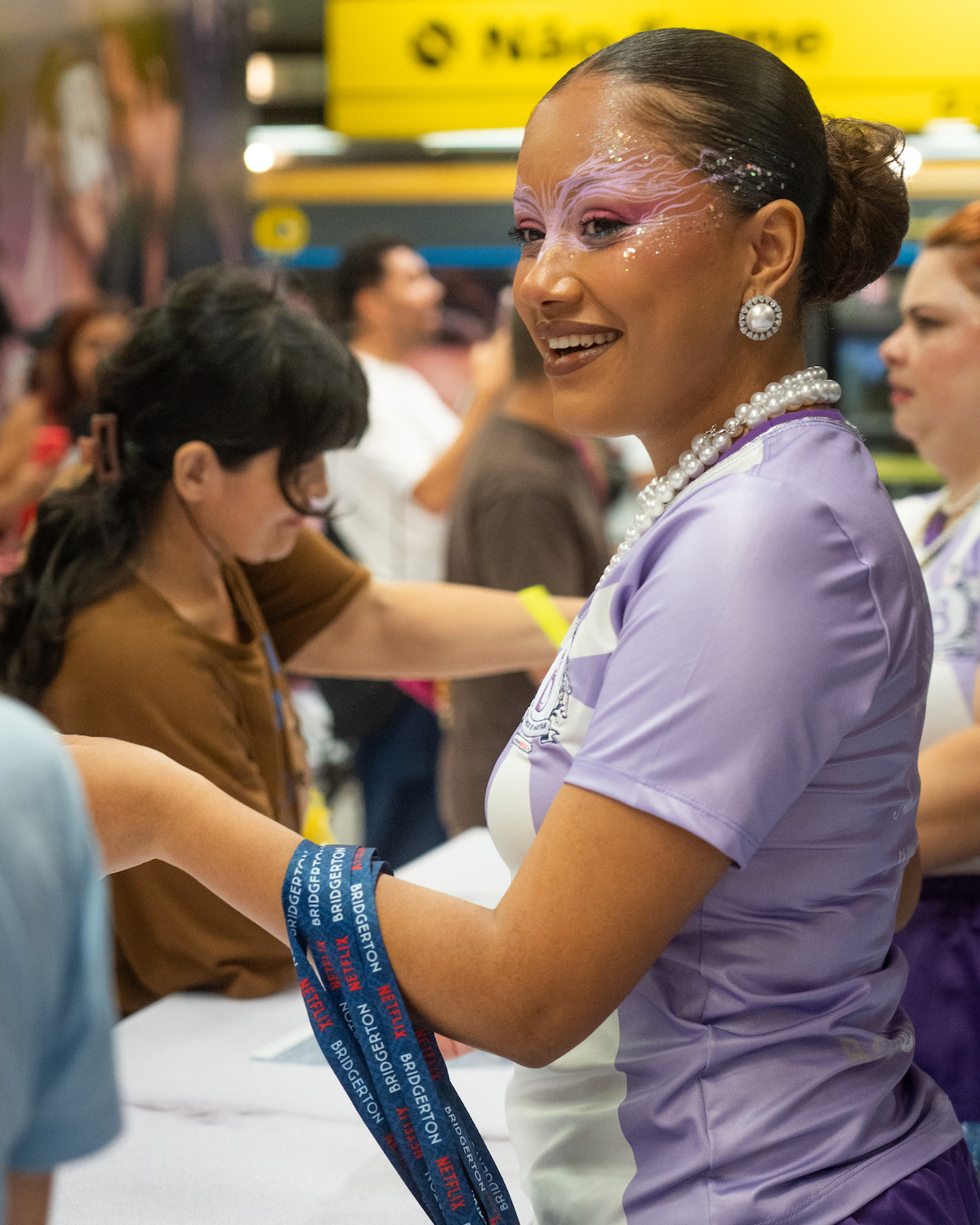Mulher sorridente, vestindo um traje lilás, maquiada com glitter e pérolas. Ela interage com pessoas em um evento movimentado em ambiente interno, com uma placa amarela ao fundo.