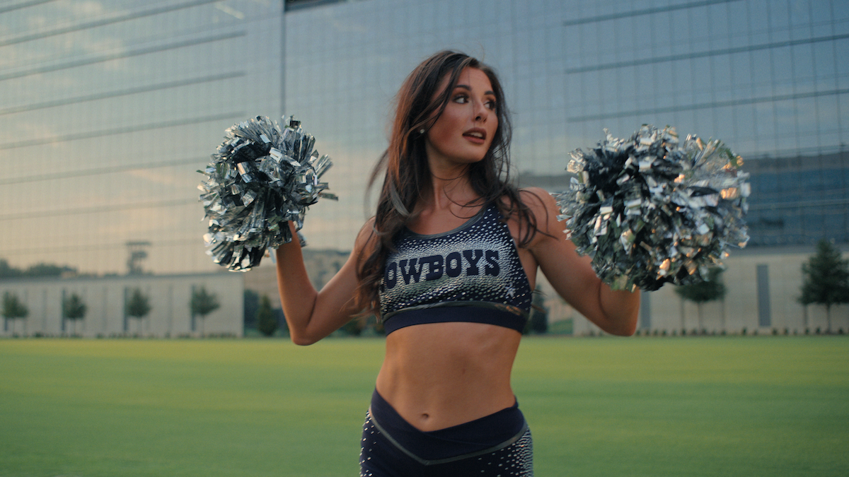 Cheerleader in a "Cowboys" uniform holding pom-poms, standing on a green field with a modern glass building in the background, looking off to the side.