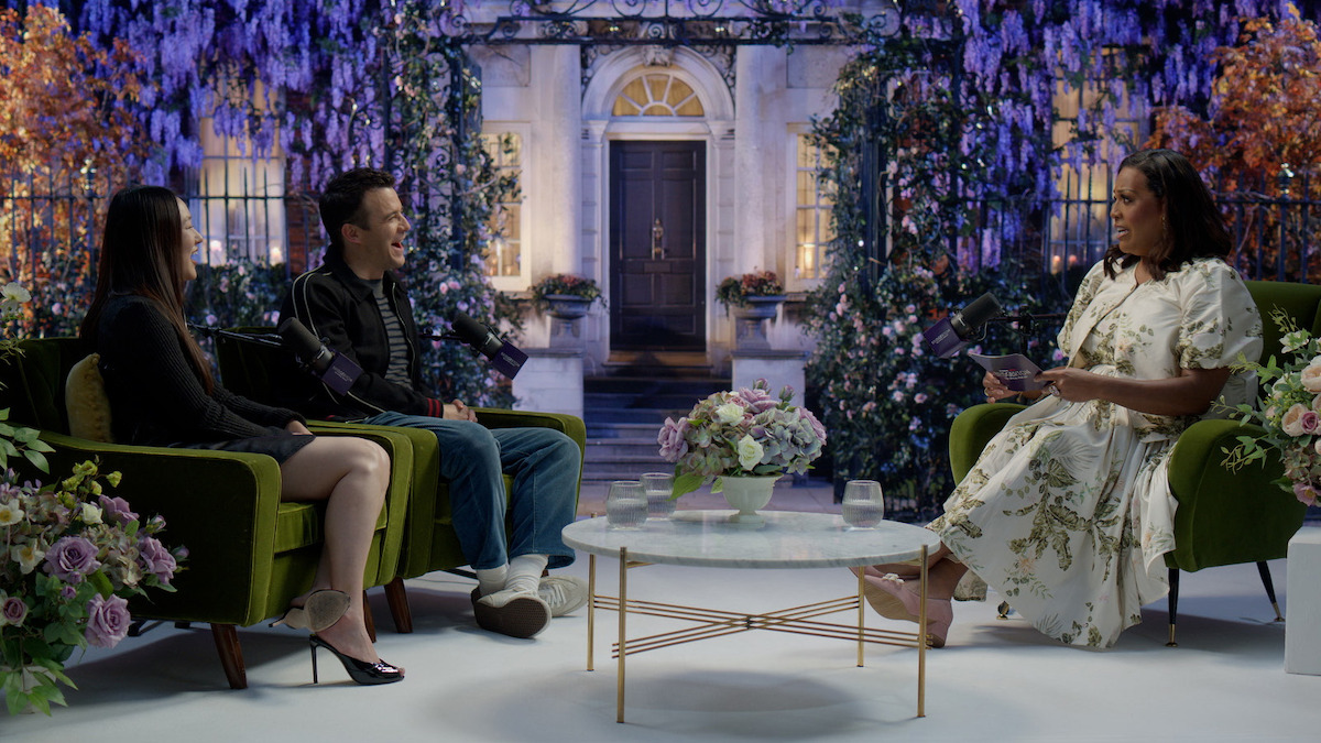 Three people sit on green chairs in a floral-themed studio set with a garden backdrop, having a conversation around a glass coffee table with floral arrangements. The setting is cozy, elegant, and brightly lit.