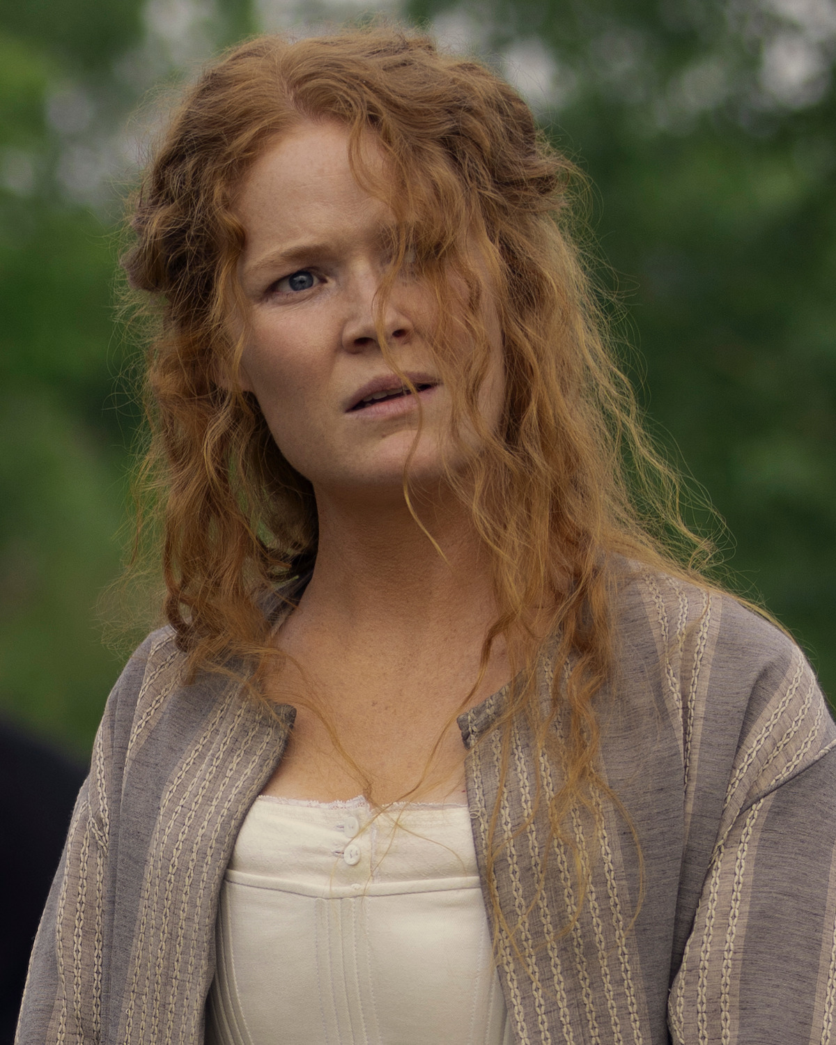 A woman with long, curly red hair in historical clothing stands outdoors, looking concerned or confused, with a blurred green natural background.