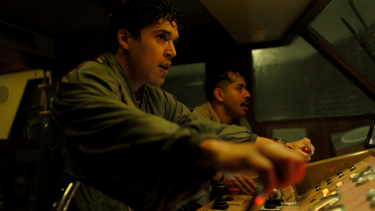 Two men in a dimly lit boat cabin operate control panels with intense focus, suggesting a tense or urgent situation at sea during stormy weather.