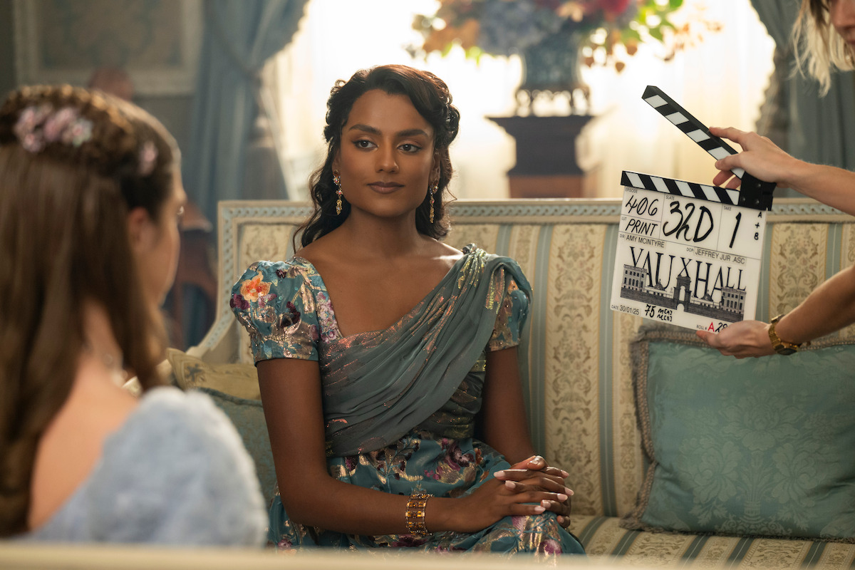 Woman in period costume sits on ornate couch in elegant, vintage room with soft light and floral decor; another woman is partially visible and a clapboard is held near the main subject, indicating a film or TV set environment.