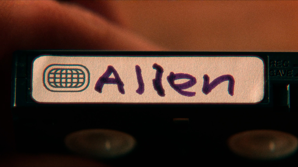 Close-up of a hand holding a labeled cassette tape with "Allen" written in marker on a white sticker, featuring a small globe-like icon on the left, in a warm indoor setting.