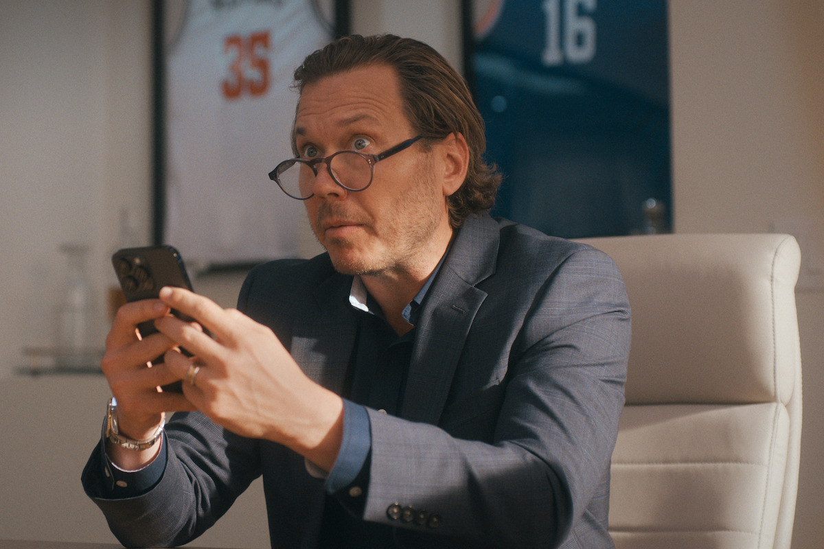 Man in a suit sitting in an office, holding a smartphone with both hands, looking focused. Framed basketball jerseys are hanging on the wall in the background.