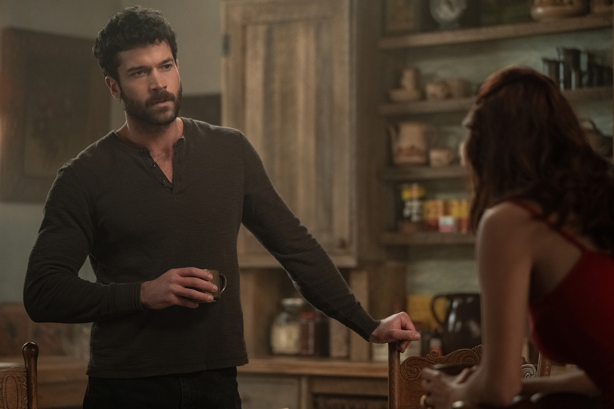 Man holding a cup stands by a wooden chair talking to a woman in a rustic kitchen with shelves holding jars and food containers, creating a cozy, homey atmosphere.