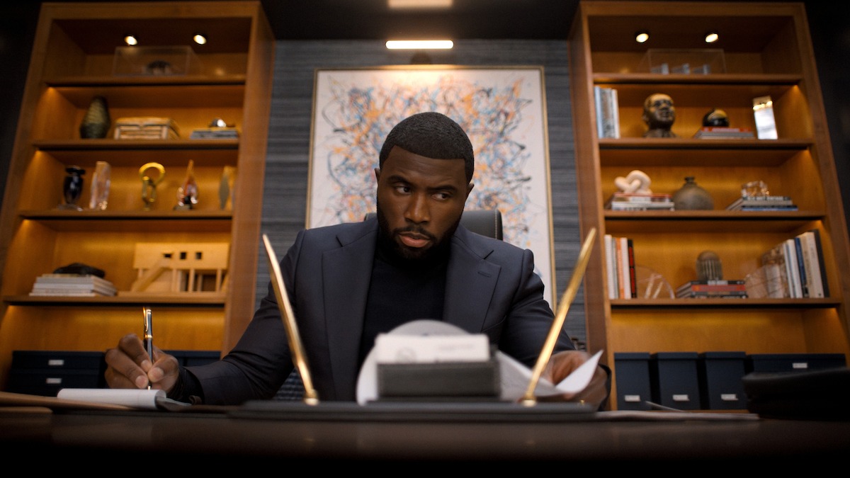 Man in a suit writing at a modern office desk, surrounded by shelves with books, art, and decor, with a colorful abstract painting on the wall behind him.