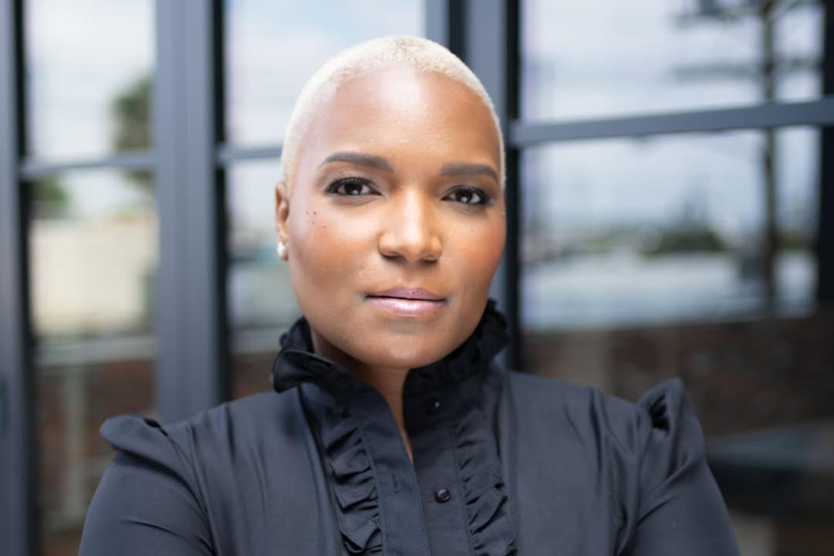 Confident person with short blond hair wearing a black ruffled shirt, standing in front of large glass windows with a blurred outdoor background.