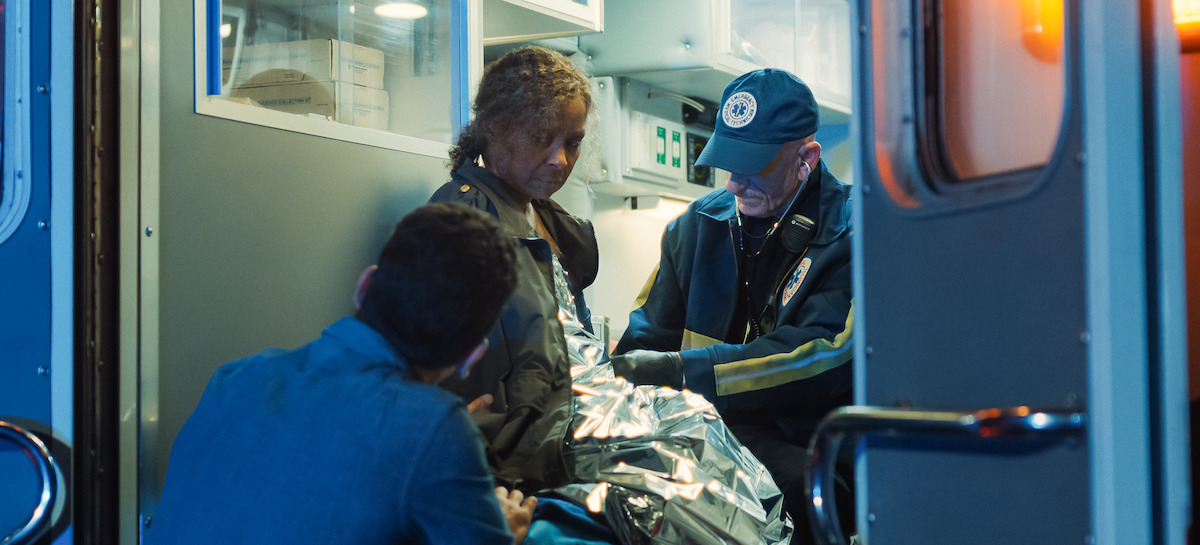 Paramedics attend to an older woman inside an ambulance at night, wrapping her in a thermal blanket.