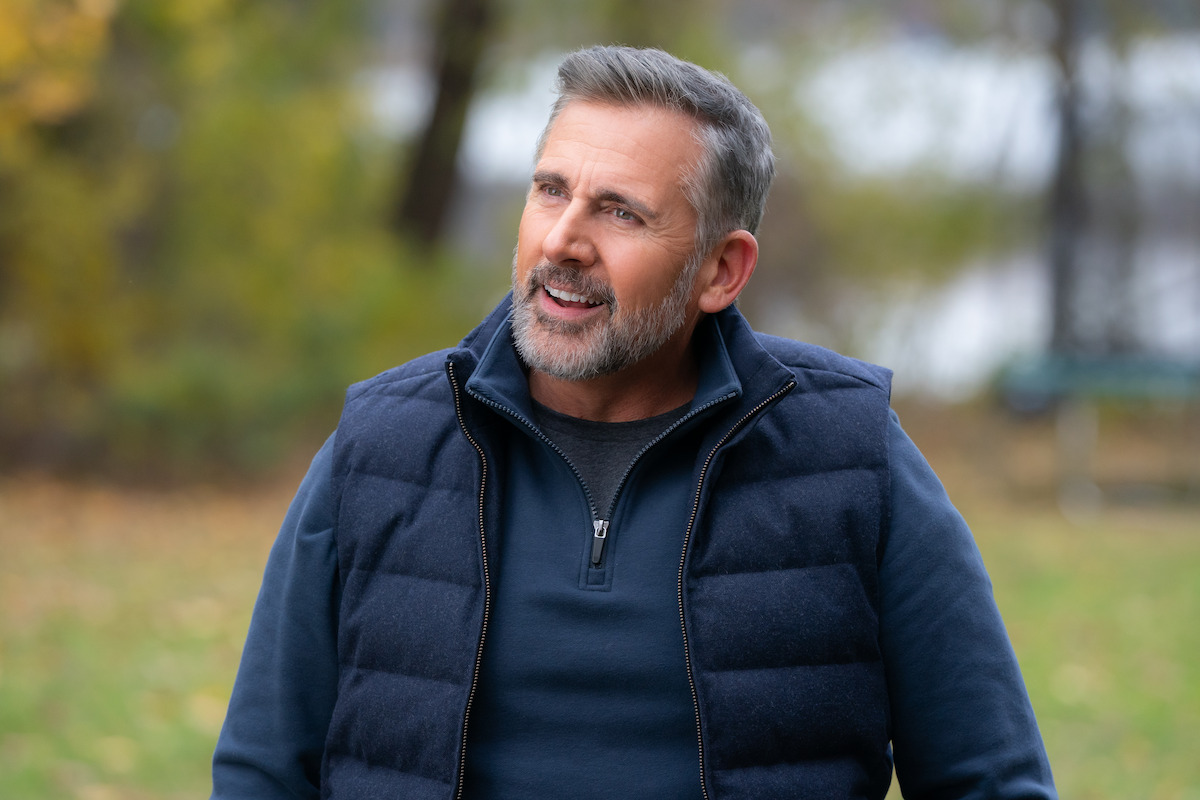 Middle-aged man with gray hair and beard wearing a navy vest and sweater, standing outdoors in a park with autumn trees and a blurred lake in the background, looking up and smiling.