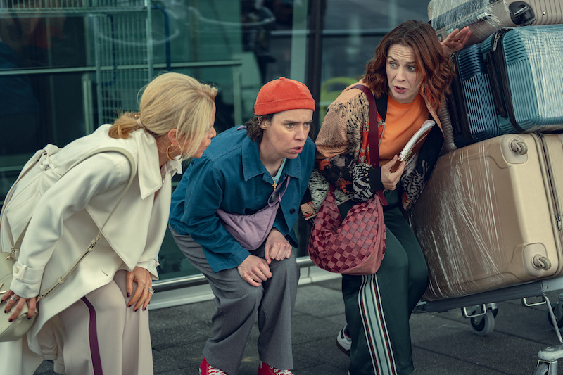 Three women squatting and talking intensely outside, next to a stack of suitcases on a luggage cart, with glass windows and an airport-like environment in the background.