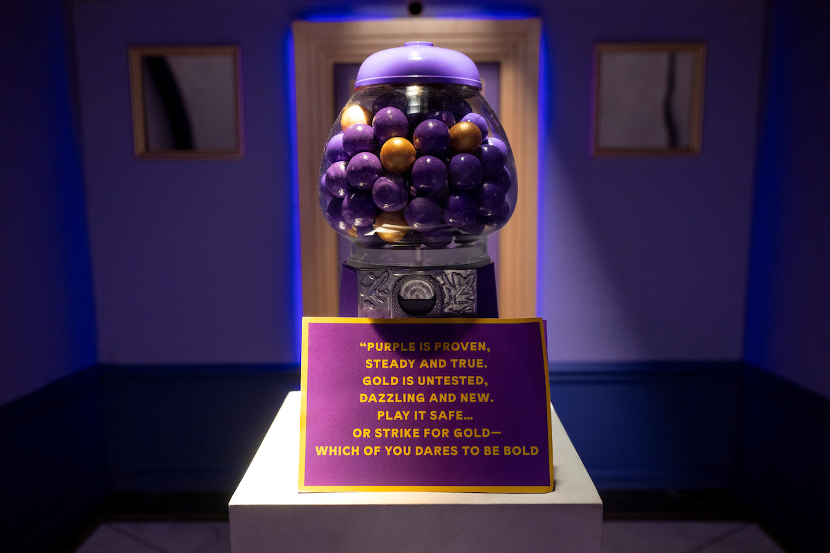 A gumball machine filled with purple and gold balls sits on a pedestal in a dimly lit room. A sign in front asks whether to play it safe with purple or take a risk with gold, inviting visitors to make a bold choice.