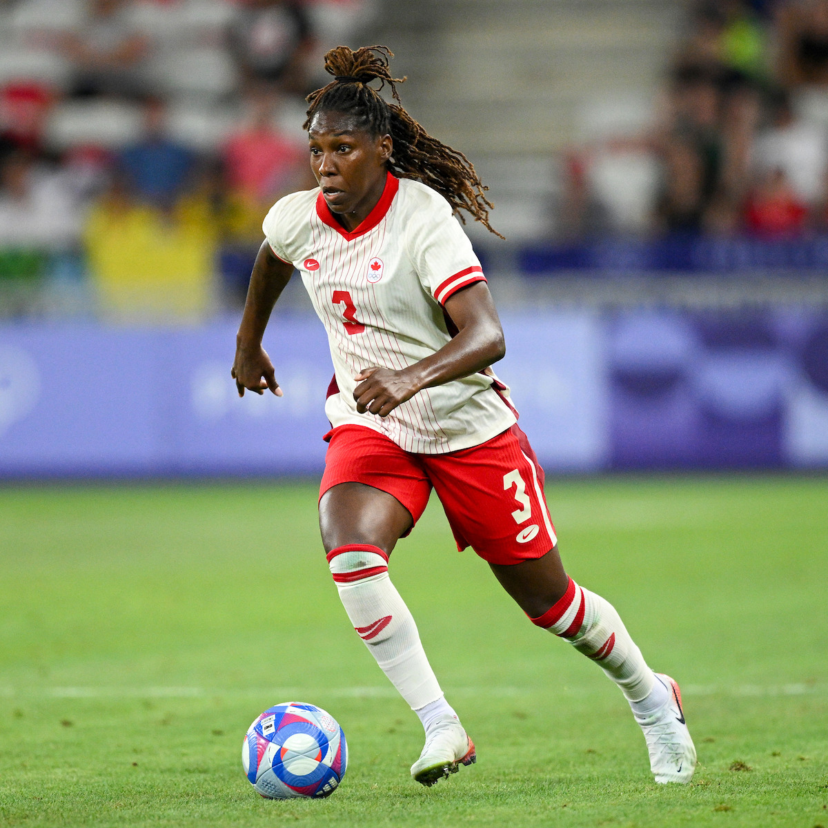 Female soccer player in a white and red uniform running with the ball on a green field during a match, with stadium seats and spectators blurred in the background.