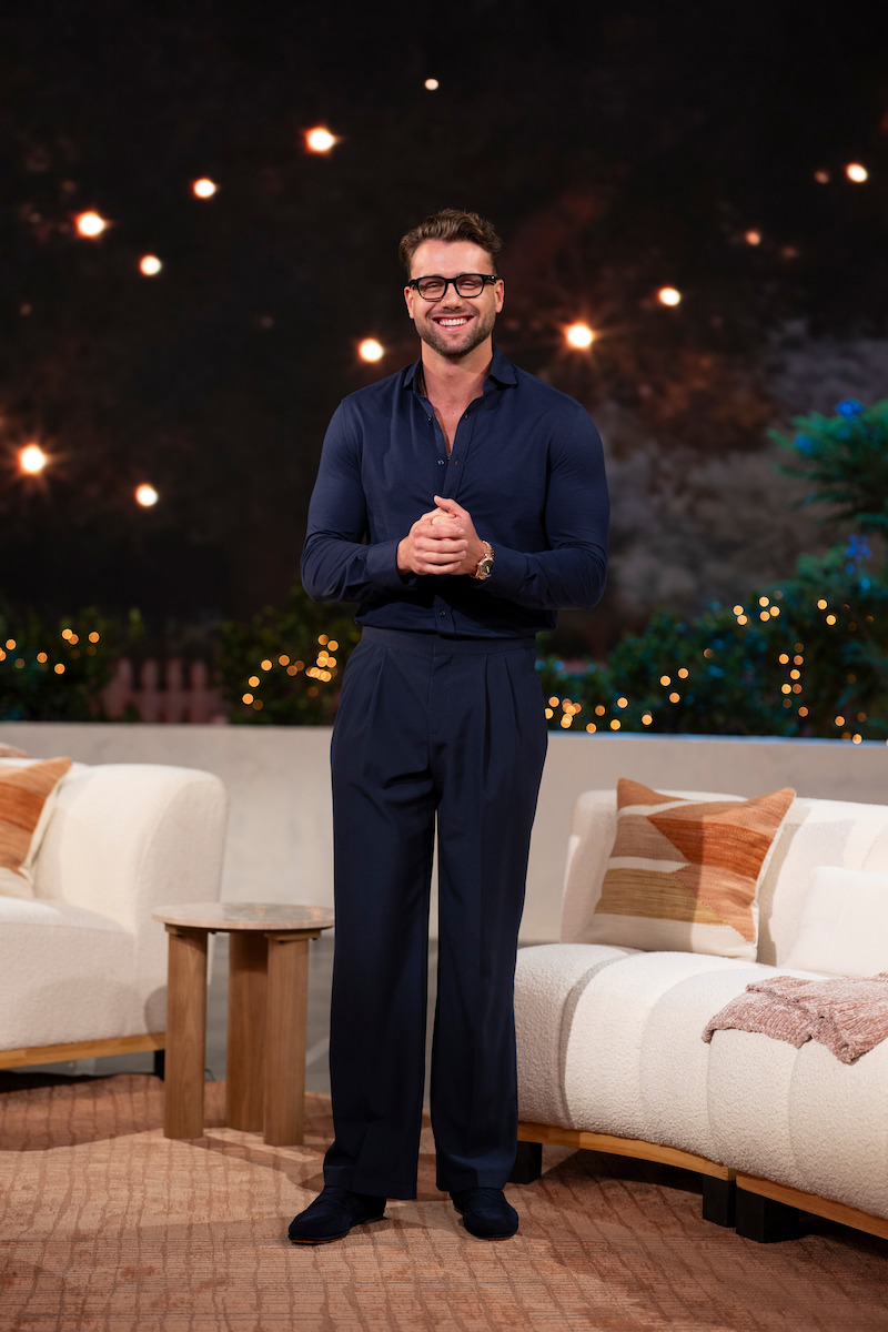 Smiling man in dark clothes stands between white couches in a cozy, warmly lit living room with string lights and greenery in the background, creating a relaxed, inviting atmosphere.