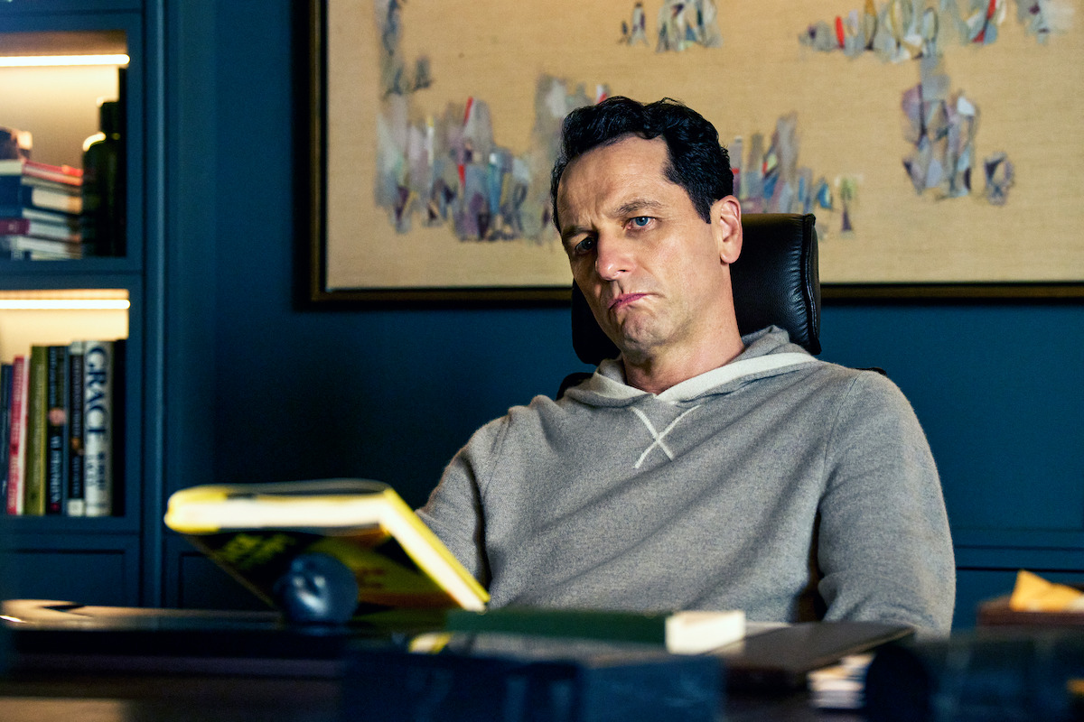 A man in a gray hoodie sits at a desk in a modern, dimly lit office, reading a yellow book with shelves and abstract art behind him, conveying a thoughtful and serious mood.