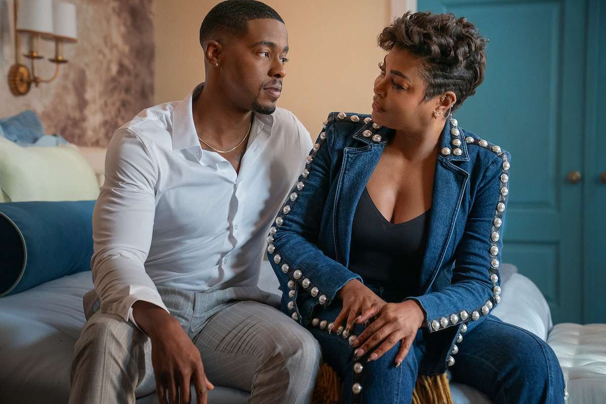 A man in a white shirt and a woman in a blue jacket with pearl accents sit closely on a bed in a well-lit, modern bedroom, looking at each other and seemingly having an intimate conversation.