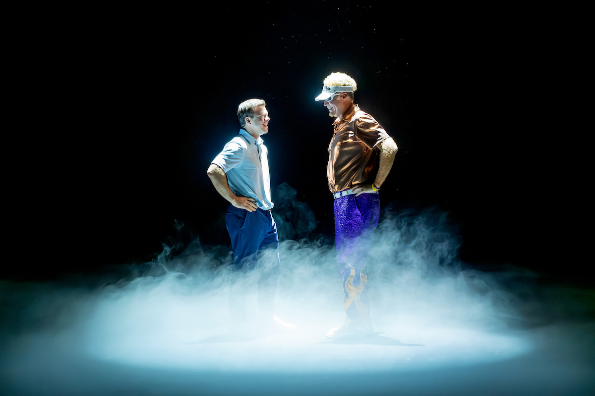 Two men stand face to face surrounded by dramatic stage smoke and spotlight, one in business attire and the other in casual sportswear, suggesting a tense or competitive scene in a dark theatrical setting.