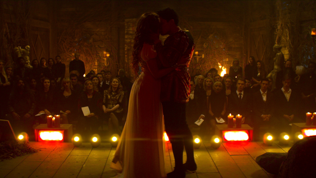 A couple embraces and kisses on a dimly lit stage, surrounded by an audience, candles, and warm yellow lighting in a dramatic, ceremonial setting.