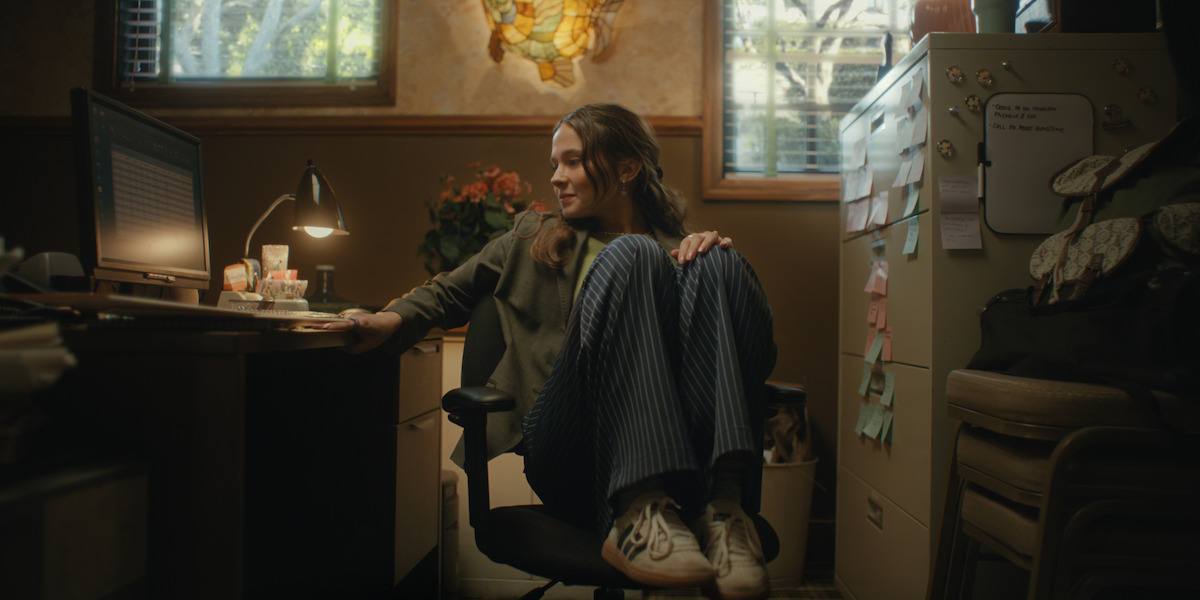 Young woman sitting in an office chair with knees up, smiling at a computer screen in a cozy, warmly-lit office with a desk lamp, file cabinet, plants, and sticky notes on the wall.