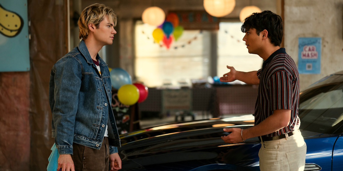 Two young men stand by a car indoors, one gesturing as if in a serious conversation. Colorful balloons and festive decorations suggest a party or event, with natural light streaming through large windows in the background.