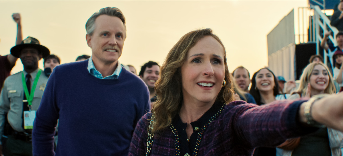 A woman and man stand outdoors in a crowd at an event. The woman is reaching forward and smiling, while people cheer and watch in the background. The atmosphere is lively with several spectators and a uniformed officer.