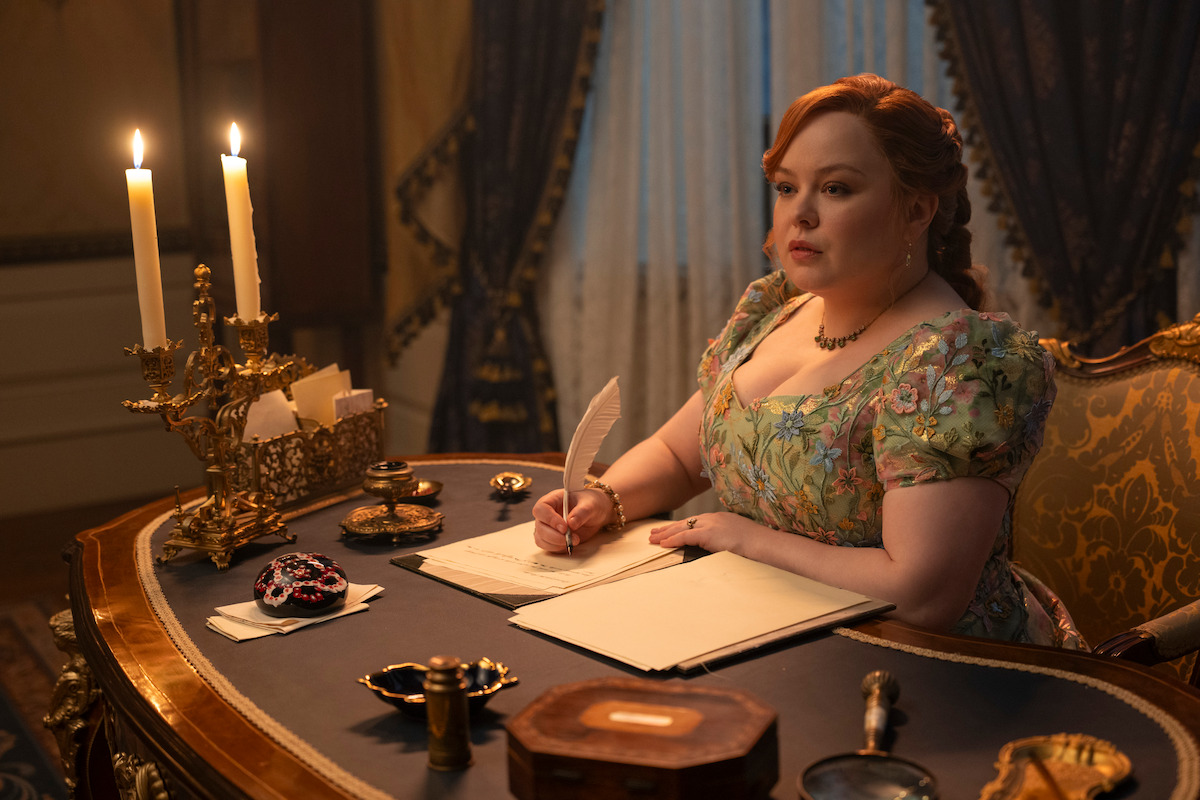 Woman in a floral dress sits at an ornate desk with a lit candelabra, writing in a notebook with a quill in an elegant, dimly lit room decorated in vintage style.