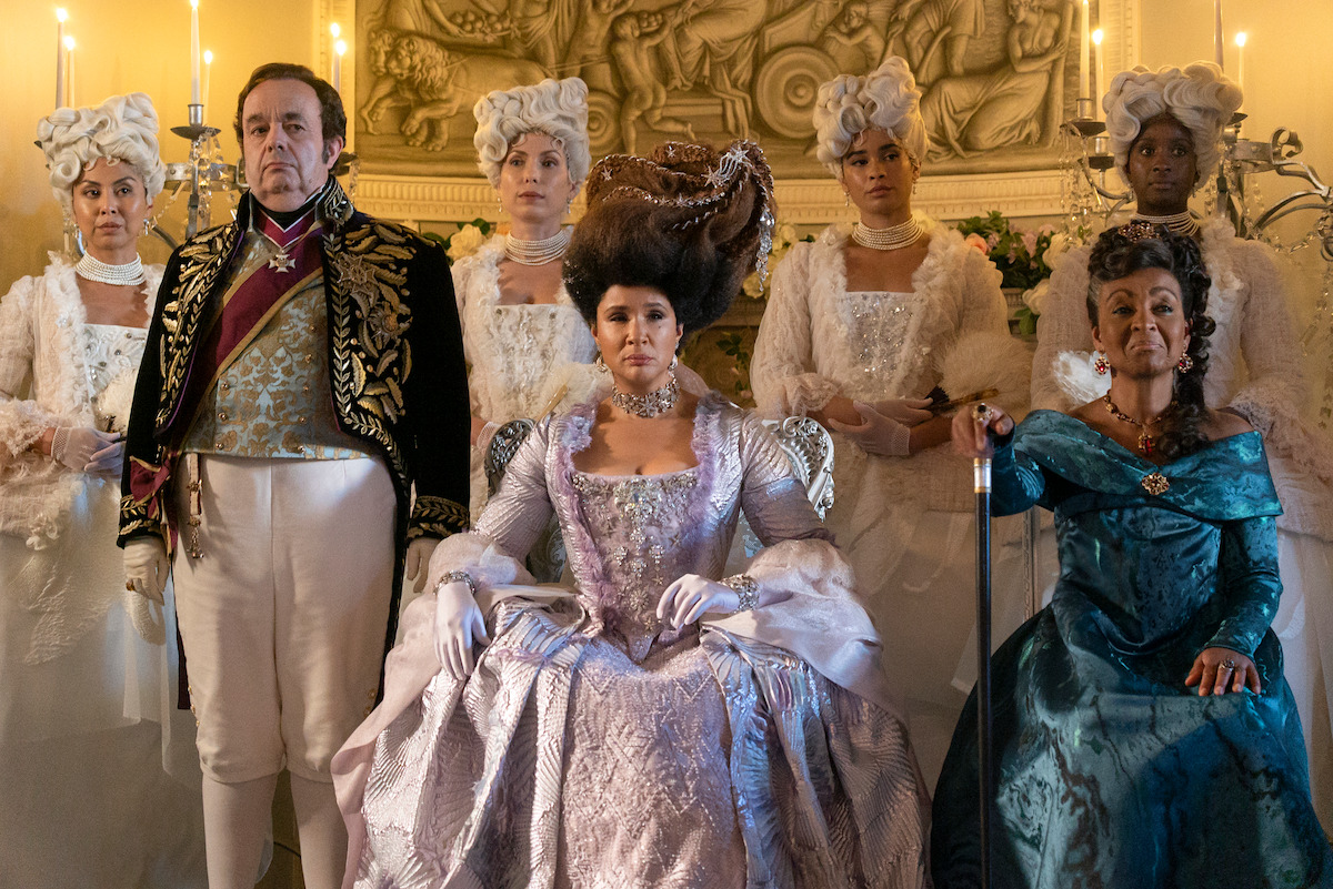 A group of people in elaborate 18th-century costumes pose formally in a lavish, candle-lit room with ornate decor and historical ambiance, highlighting regality and period drama.