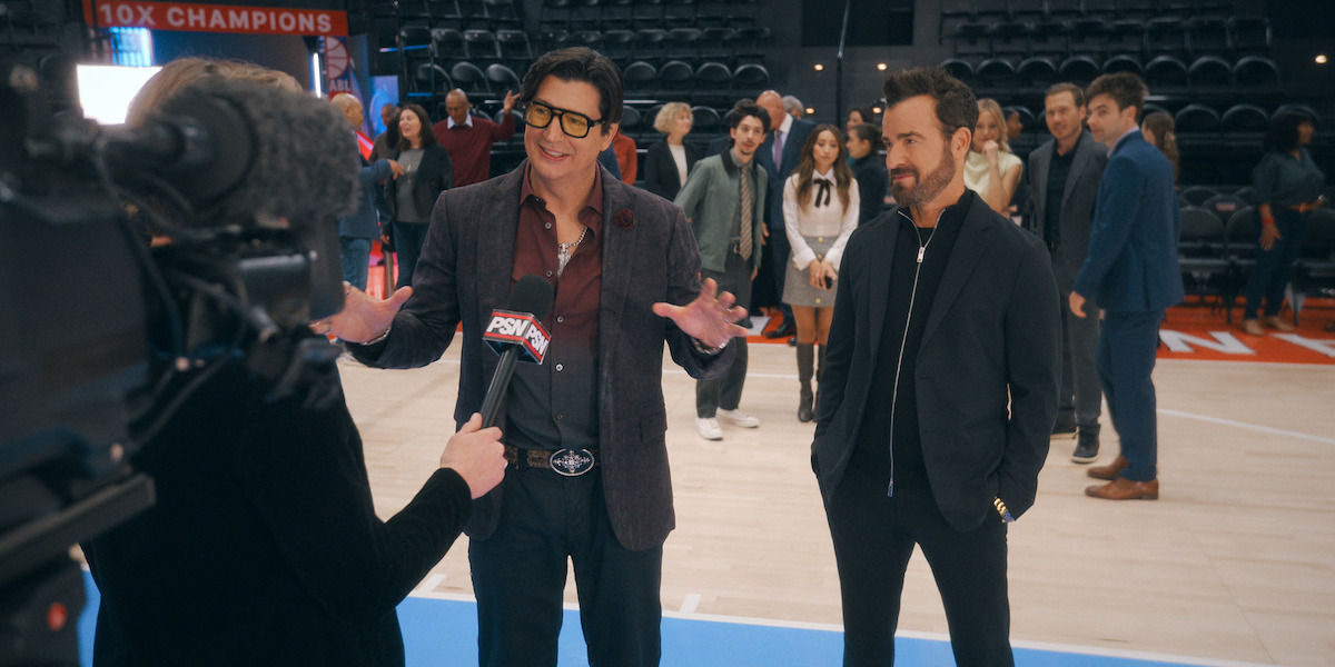 Two men are being interviewed by a news reporter on a basketball court, with a camera filming them. In the background, a group of people stand watching in an arena setting.
