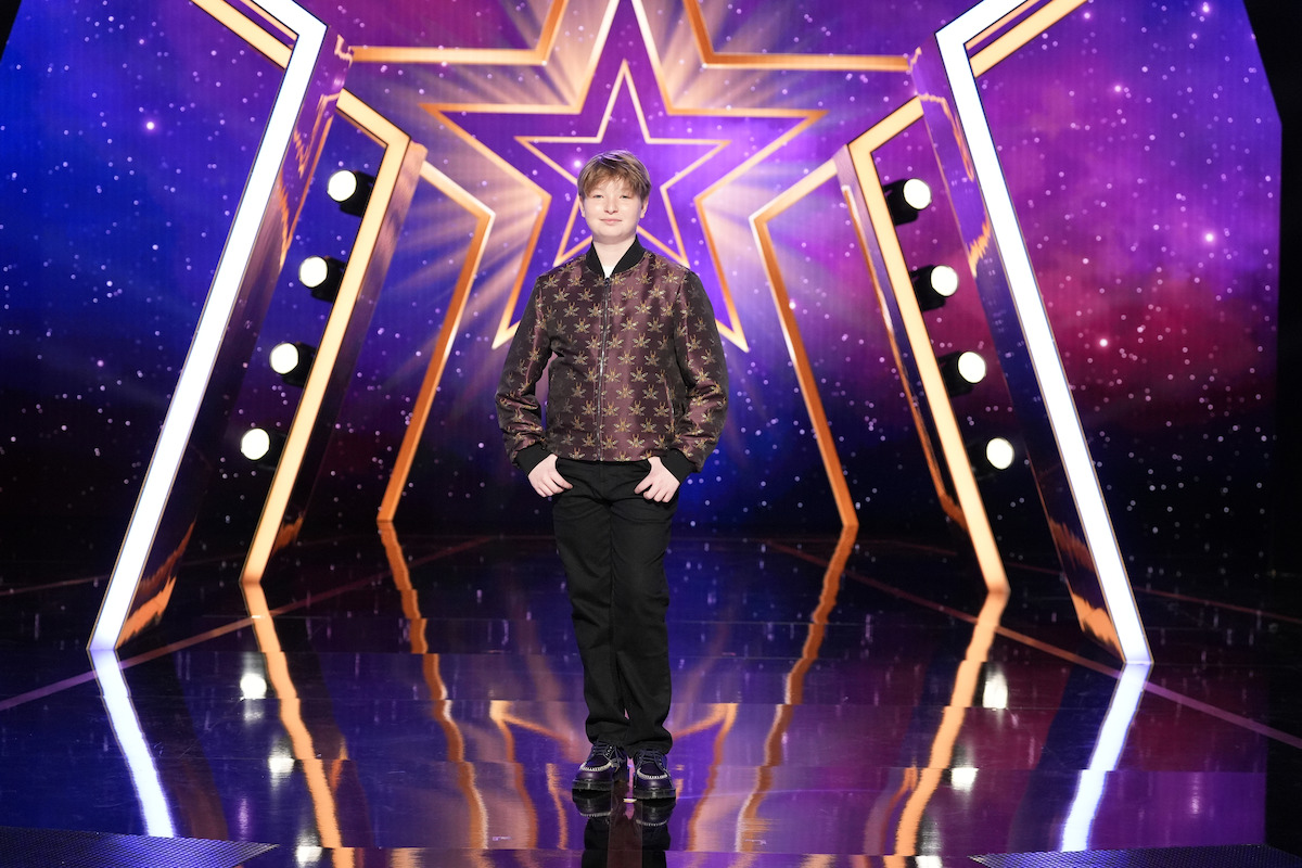 Yair Keydar se encuentra en un escenario brillante con un fondo estrellado de color púrpura, estructuras geométricas doradas y luces brillantes del escenario, creando un escenario dramático y vibrante para un espectáculo de talentos.