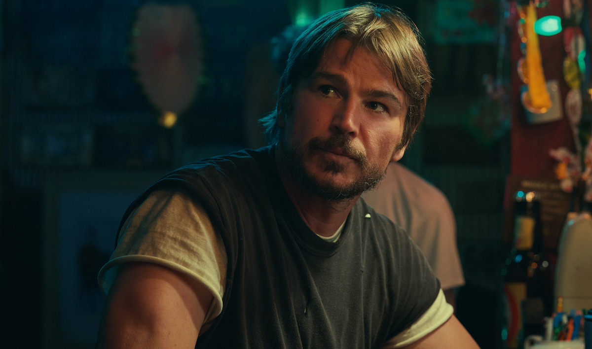 Man with light brown hair and beard sitting at a dimly lit bar counter, wearing a dark t-shirt over a white undershirt, with colorful decorations and bottles visible in the background.