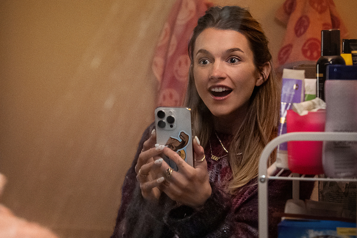 A young woman with long hair smiles excitedly at her phone in a bathroom. Shelves with toiletries and a pink towel are visible in the warm, softly lit environment.