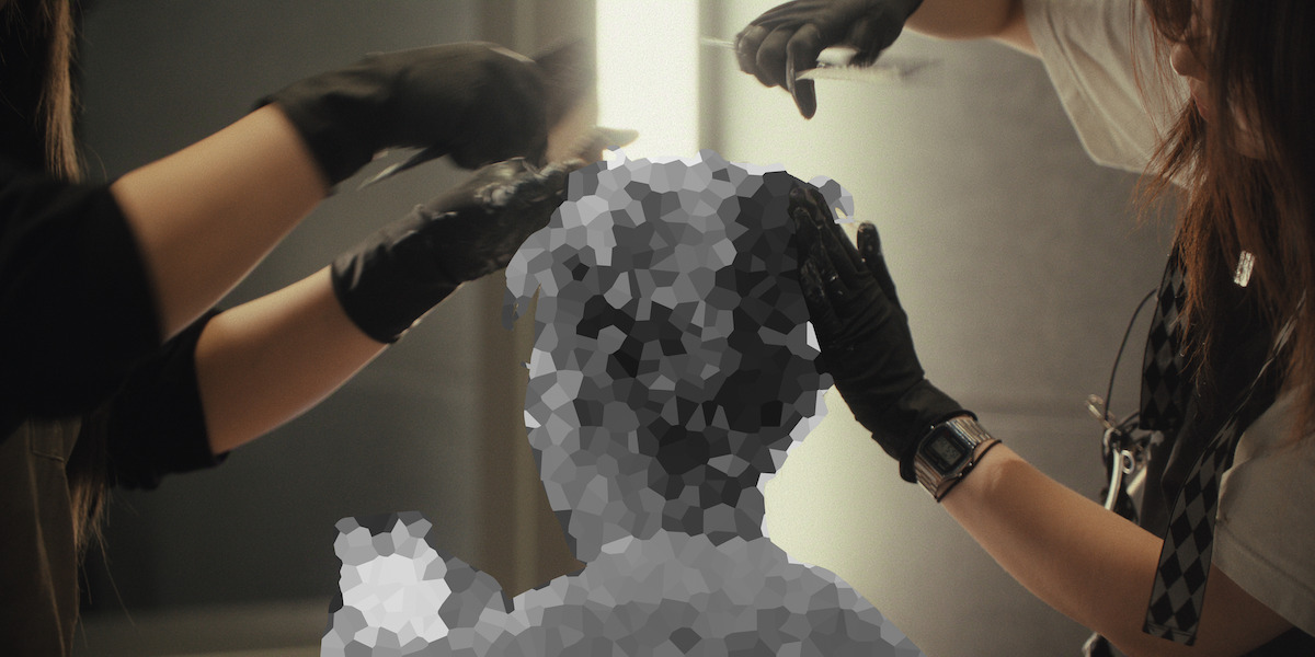 Two people wearing black gloves are styling or cutting a person’s hair in a salon-like setting, but the client’s head and upper body are heavily pixelated, obscuring their features.