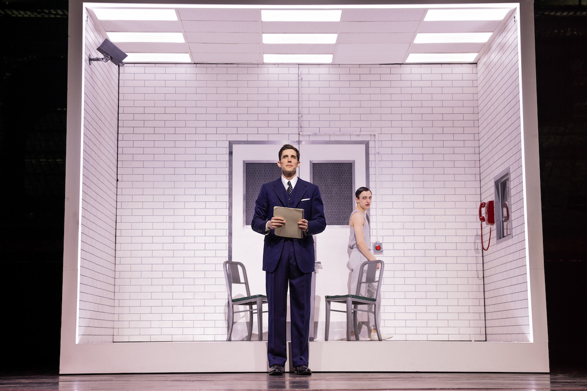 A man in a suit holds a clipboard standing in a brightly lit, white-tiled room with a woman seated behind him, a table and chairs, a red wall phone, and a surveillance camera, resembling an interrogation or observation room.