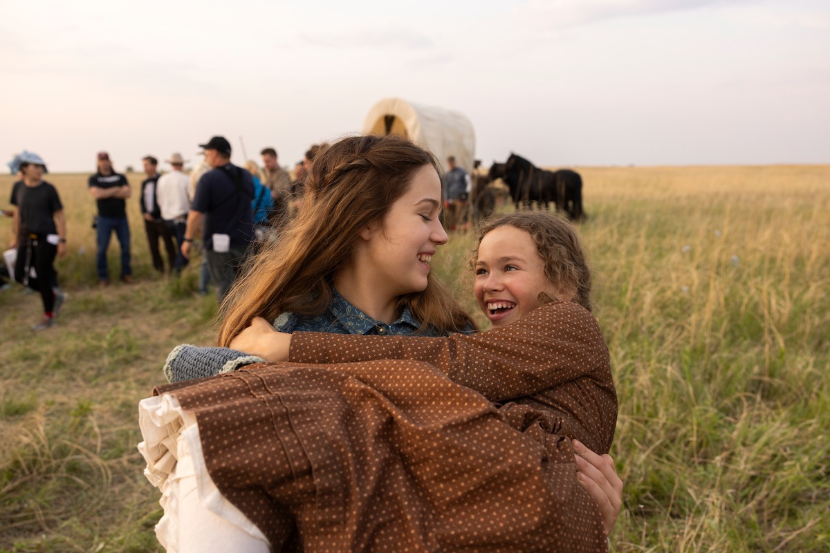 Two girls in pioneer-style clothing smile and embrace outdoors in a grassy field, with a group of people, horses, and a covered wagon in the background under a partly cloudy sky.