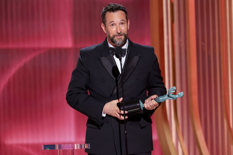 Noah Wyle in a black tuxedo holding an award and speaking at a microphone on stage with pink and gold background, spotlighted during an awards ceremony.