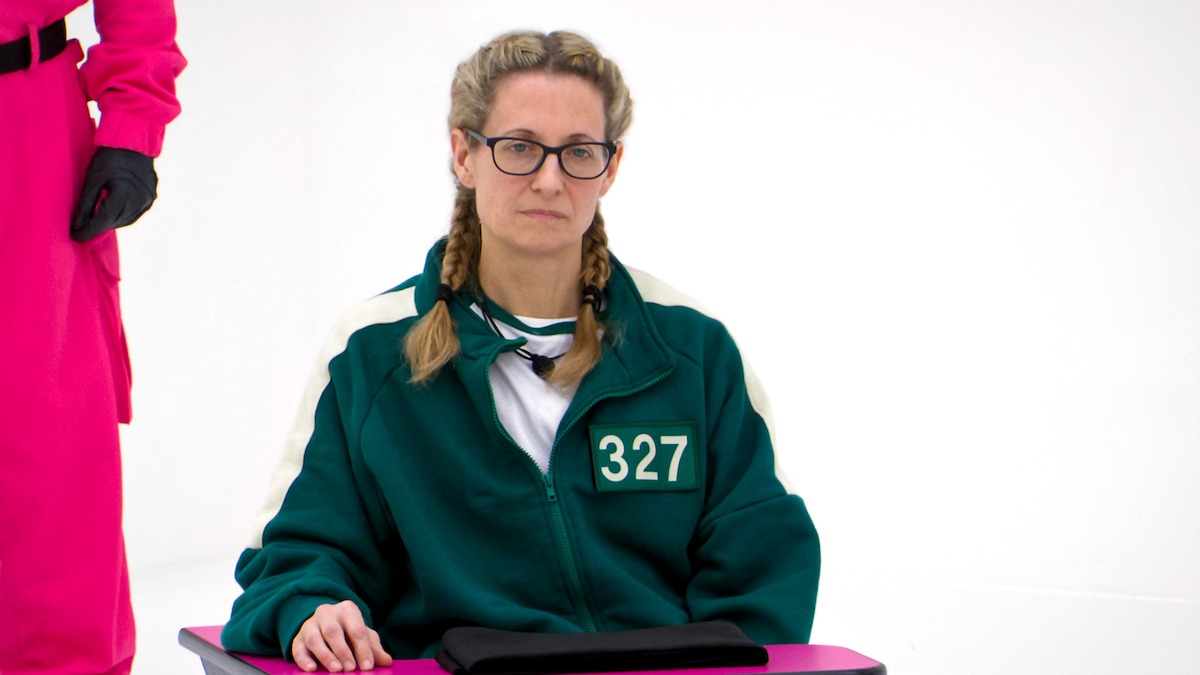 A woman with glasses and braided hair sits at a small table in a green tracksuit marked 327, in a bright, stark white room. The mood is tense and serious; another person in a pink uniform stands nearby.