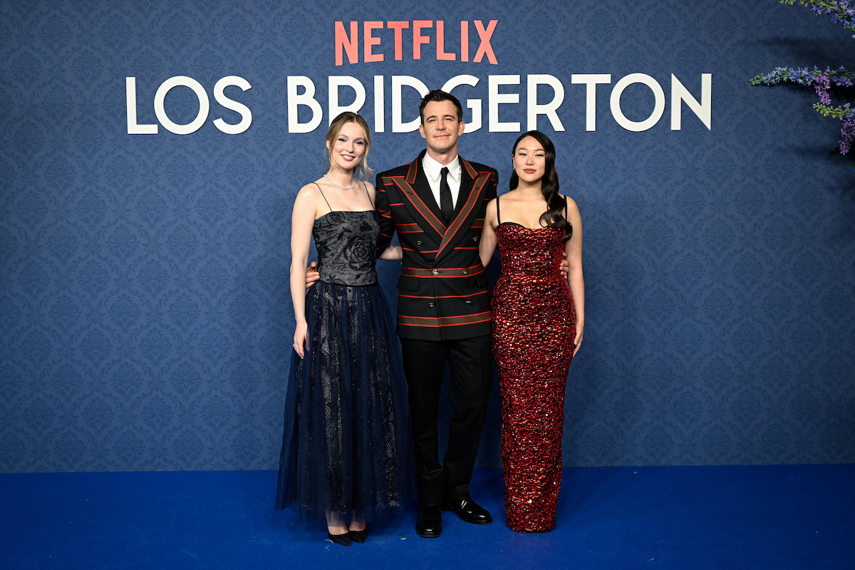 Três pessoas usando trajes formais posam em um tapete azul durante um evento de ‘Bridgerton’, da Netflix, com um painel azul estampado e a sinalização do evento ao fundo.