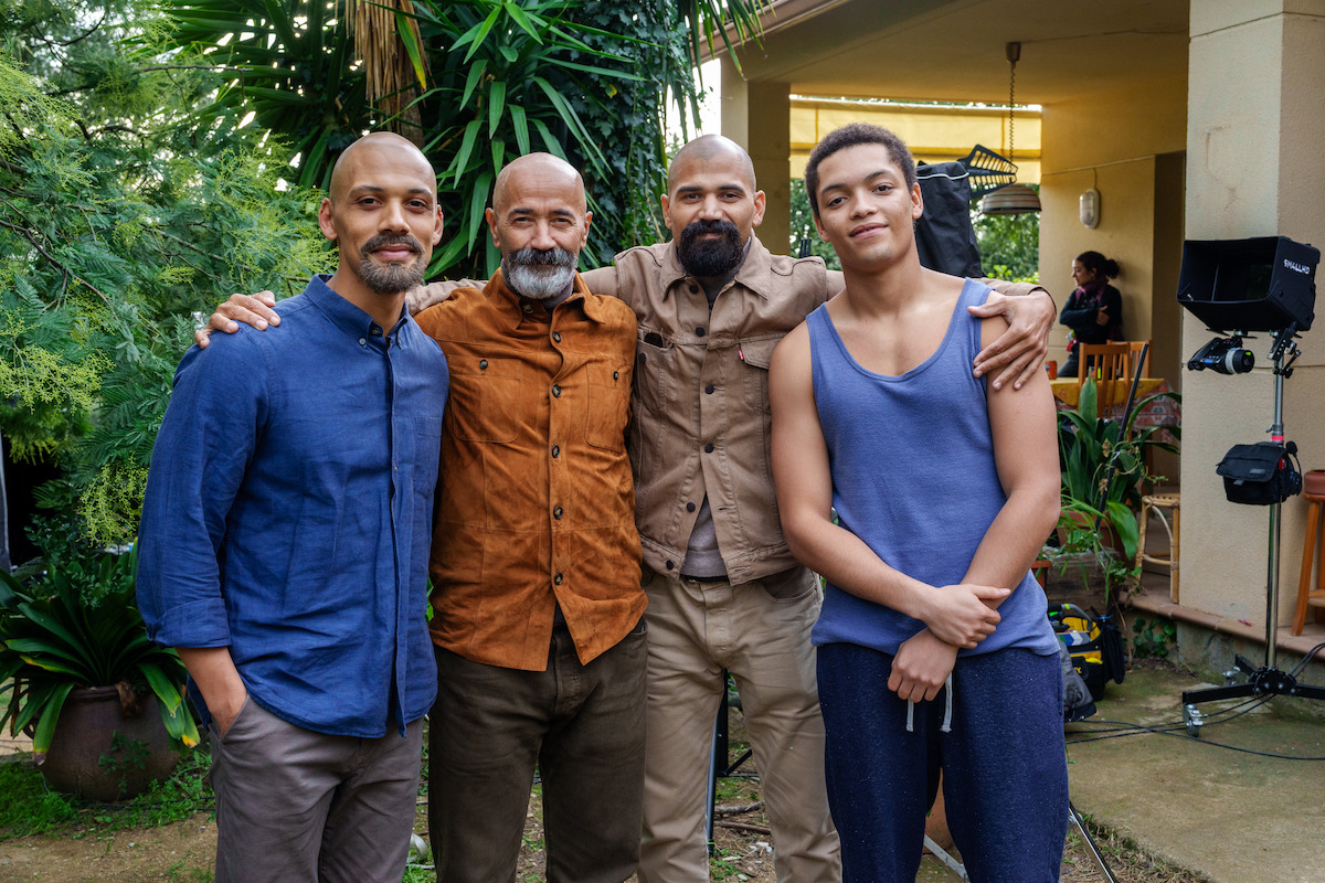 Four men standing together outdoors in a garden near a house, smiling and posing for a group photo, with lush green plants and film equipment visible in the background.