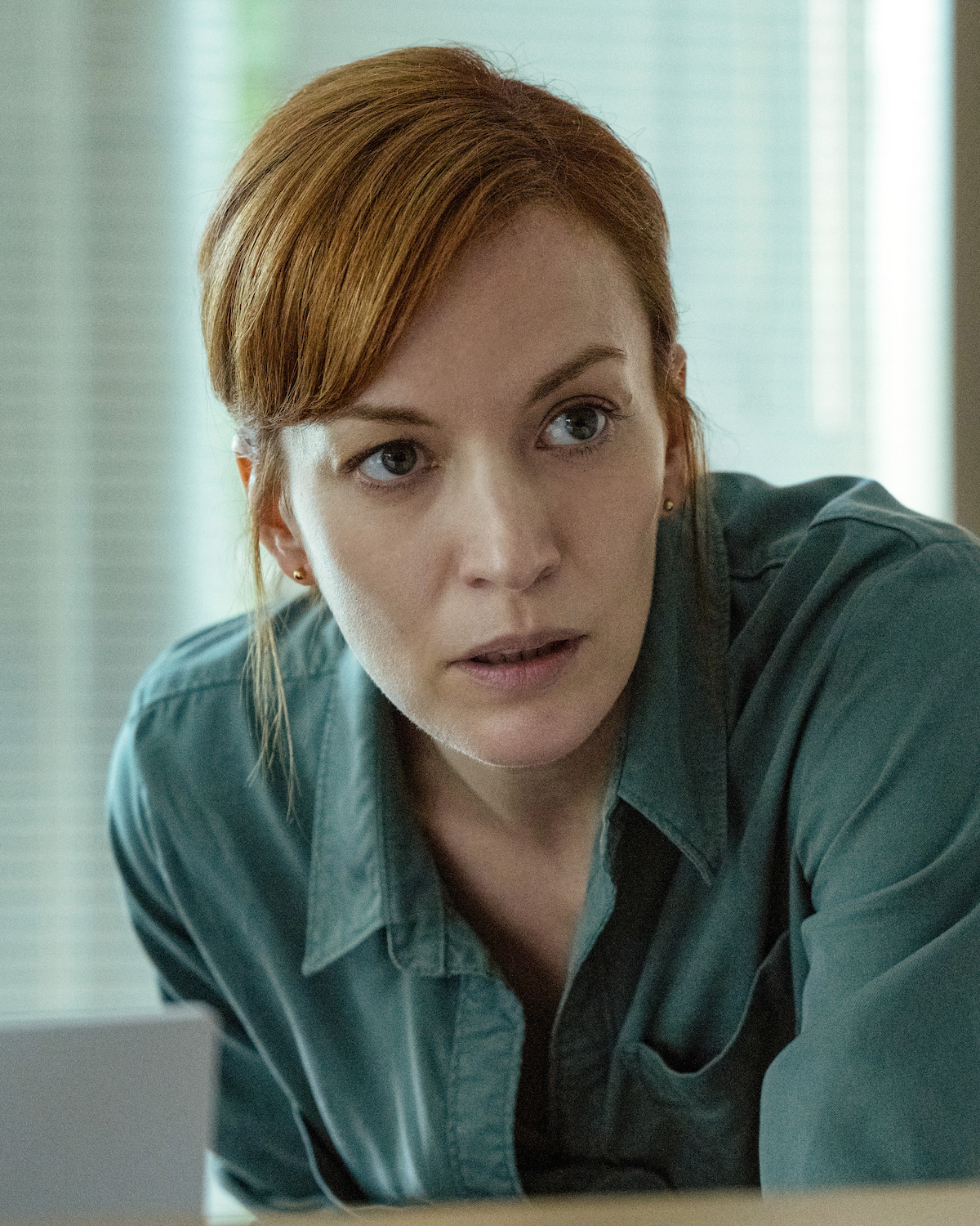 Woman with red hair in a green shirt leaning forward in an indoor setting with natural light and blurred background featuring blinds and window.
