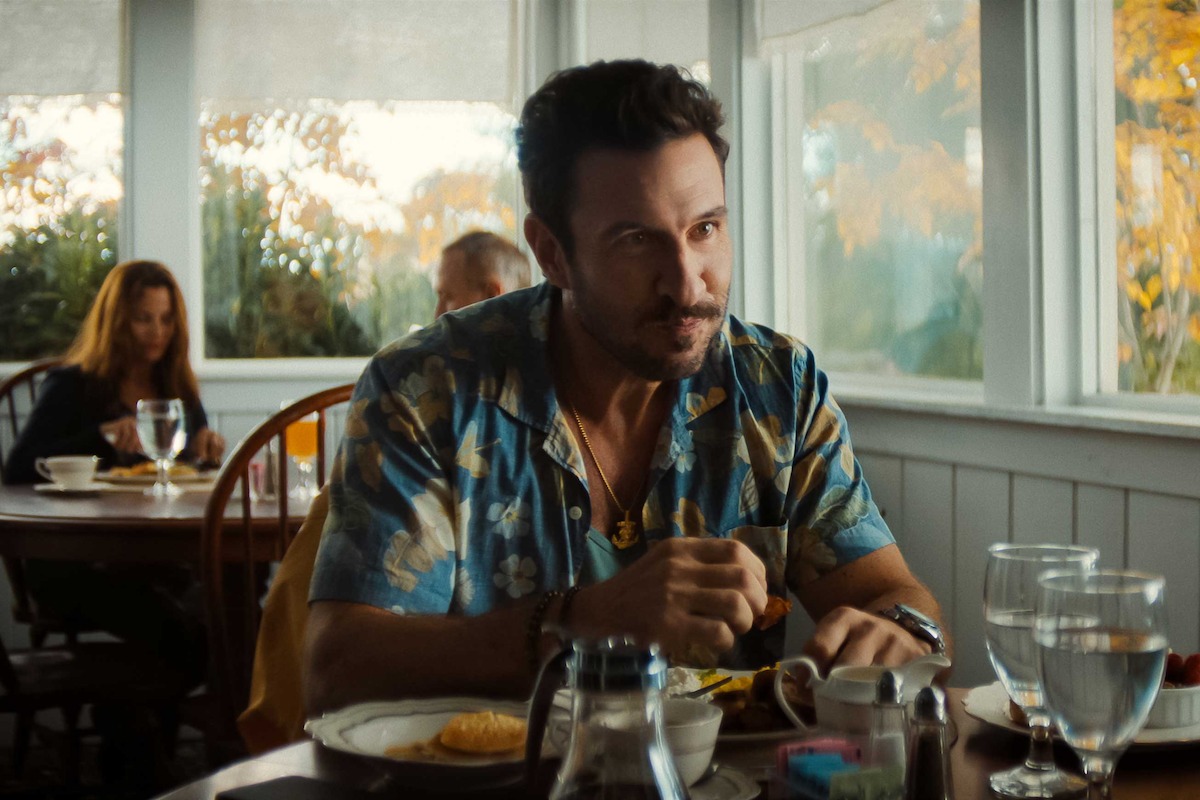 Man in a floral shirt eating breakfast at a sunlit diner table, with food, coffee, and utensils on the table; other patrons sit and eat in the background near large windows showing autumn trees.